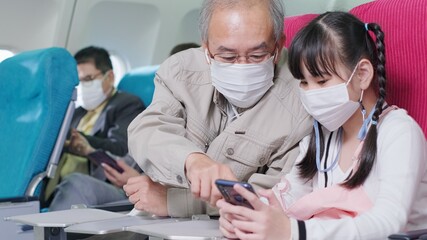 Asian grandfather is teaching to wear correctly the facial mask to his granddaughter.Coronavirus prevention on the airplane.travel concept