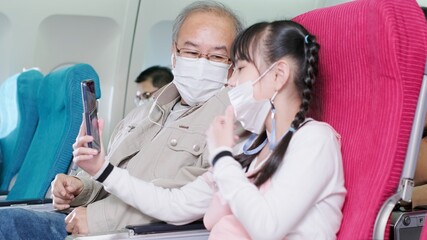 Asian grandfather is teaching to wear correctly the facial mask to his granddaughter.Coronavirus prevention on the airplane.travel concept