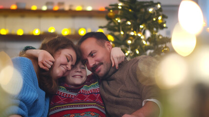 Happy mom, dad and preteen son embracing relaxing on couch in apartment decorated for christmas