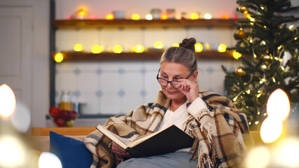 Senior woman sitting on sofa with book in room with christmas decorations.
