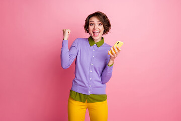 Photo of excited lady hold telephone raise fist lucky day wear purple jumper yellow pants isolated pink color background