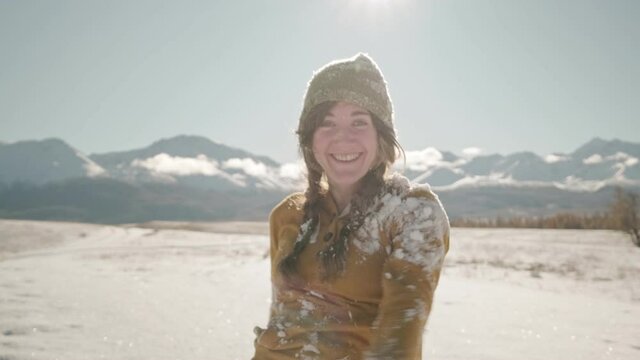 Portrait of a cheerful woman in a sweater and a hat with a pom-pom throwing snow up. Snowflakes sparkling in the sun falling down