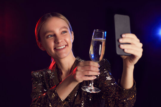 Portrait of blonde young woman taking selfie photo via smartphone while enjoying party in night club and toasting with champagne glass