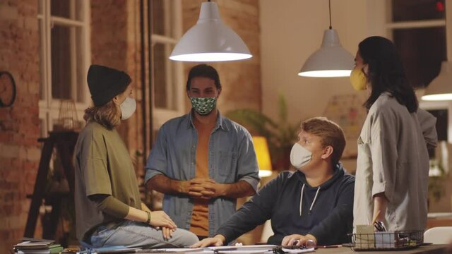 Diverse team of young male and female colleagues in casualwear and protective masks discussing business while working late in modern loft office during coronavirus pandemic