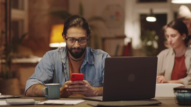 Tilt down shot of handsome middle eastern man sitting at desk and messaging on smartphone while working late with colleagues in loft office