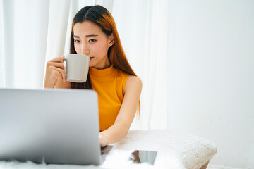 Asian woman drinking cup of water and using laptop computer at home.