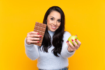 Young Colombian girl over isolated yellow background taking a chocolate tablet in one hand and an...