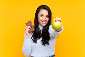 Young Colombian girl over isolated yellow background taking a chocolate tablet in one hand and an...