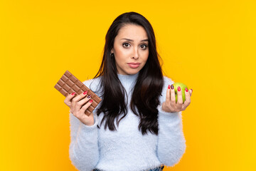 Young Colombian girl over isolated yellow background taking a chocolate tablet in one hand and an apple in the other