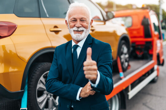 Elegant senior businesman is happy and satisfied with fast and reliable towing service for help on the road. He is standing in front of wrecker with arms crossed. Roadside assistance concept.