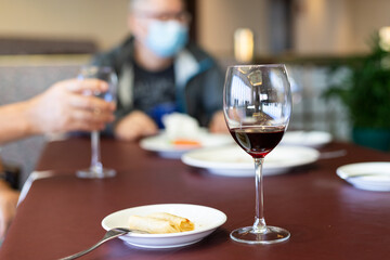 wine Glass and spring rolls on table, blurred person with face mask in background. Gathering in bar or restaurant limited to 4 people, leading to bad business. face mask mandatory in public settings