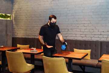 Waiter wearing protective face mask and gloves while disinfecting tables indoor restaurant, cafe. Precautions during the covid-19 coronavirus pandemic