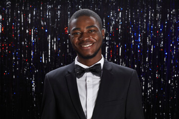 Portrait of smiling African-American man wearing tux posing at party against sprkling background,...