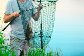 The Caucasian fisherman is standing near the water in the grass and holding the fishing rod and the...