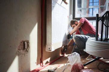 Side view of male heating engineer in work overalls using electric drill while laying pipes on the...