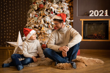 Father and son in christmas hats are enjoying near christmas tree in modern decorated home, Happy...