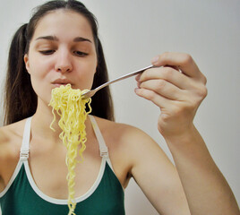 Young girl with dark hair eating instant noodles with a fork on light background