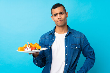 Young handsome man holding waffles over isolated blue background with sad expression