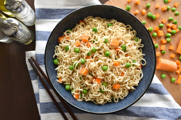 Instant noodles with vegetables (carrot and peas) and boiled egg on a blue bowl with chopsticks.