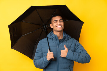 Young handsome man holding an umbrella over isolated yellow background with thumbs up because...