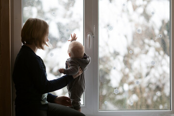 Young woman and her small child sitting on the window sill. Window is decorated for New Year. Snowy forest is outside. Christmas eve and family time concept