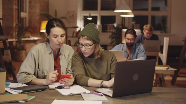 Two young women in casual outfit using laptop and smartphone while discussing project at desk in evening in open space loft office