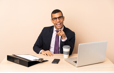 Young business man in his office with a laptop and other documents surprised and pointing front