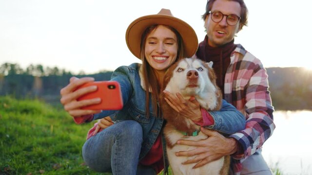 Smiling happy couple with a dog taking selfie photo on phone in autumn forest near lake. Landscape vacation relationship pets. Outdoors nature. Slow motion