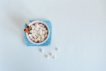 Light blue handmade knitted coaster, cup with marshmallows and cinnamon over white