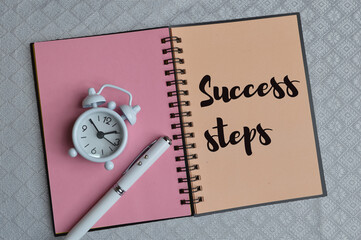 Top view of clock, pen and notebook written with text SUCCESS STEPS. Business and education concept.