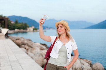 Girl traveler takes selfie with amazing view of sea, mountains, coast. Woman tourist in hat, casual clothes is using mobile phone and smiling on embankment. Travel, vacation, female tourism concept.