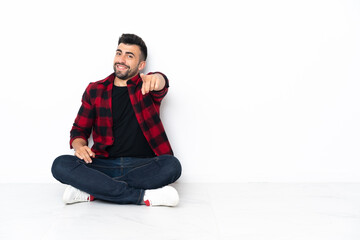 Young handsome man sitting on the floor points finger at you with a confident expression