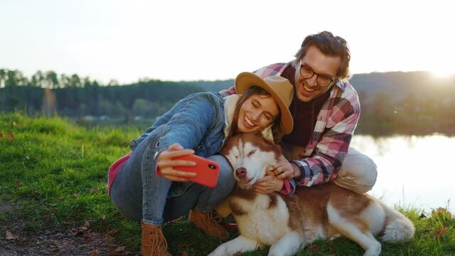 At sunlight happy couple with a dog taking selfie photo on phone in autumn forest near lake. Landscape vacation relationship pets. Outdoors nature. Slow motion