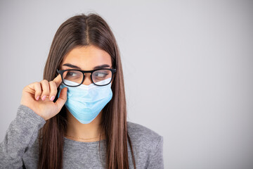 Woman wiping foggy glasses caused by wearing disposable mask on gray background, space for text. Protective measure during coronavirus pandemic