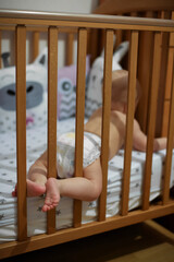 Fototapeta na wymiar The baby is lying on her stomach in the crib, her legs sticking out through the bars