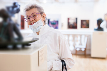 Fototapeta premium elderly European woman in mask protecting against covid examines paintings on display in hall of art museum
