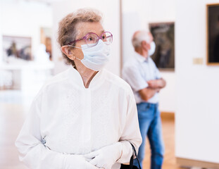 elderly European woman  in mask protecting against covid examines paintings on display in hall of...