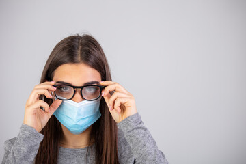 Woman wiping foggy glasses caused by wearing medical mask indoors, closeup. Woman wiping foggy glasses caused by wearing disposable mask on gray background, closeup.