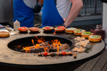 Chef preparing fish burgers with seafood, avocado, shrimp, prawn on brazier with hot flame at summer local food market - close up view. Outdoor cooking, gastronomy, cookery, street food concept