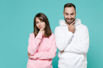 Pensive young couple two friends man woman 20s wearing white pink casual hoodie standing put hand prop up on chin looking camera isolated on blue turquoise colour wall background studio portrait.