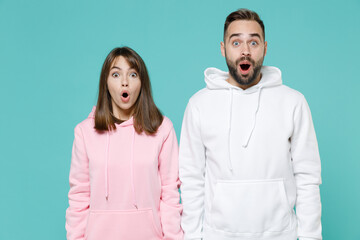 Shocked amazed young couple two friends man woman 20s wearing white pink casual hoodie standing keeping mouth open looking camera isolated on blue turquoise colour wall background studio portrait.