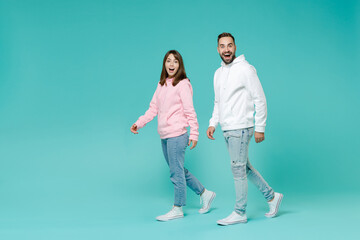 Full length side view of surprised excited young couple two friends man woman 20s wearing white pink casual hoodie walking going looking camera isolated on blue turquoise background studio portrait.