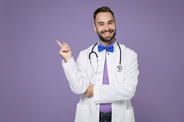 Smiling young bearded doctor man wearing white medical gown pointing index finger up on mock up copy space isolated on violet background studio portrait. Healthcare personnel health medicine concept.