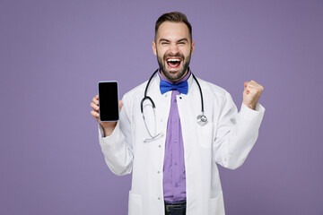 Happy young bearded doctor man in medical gown stethoscope hold mobile phone with blank empty screen doing winner gesture isolated on violet background. Healthcare personnel health medicine concept.