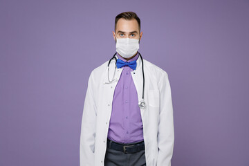 Young bearded doctor man wearing white medical gown stethoscope sterile face mask to safe from coronavirus virus covid-19 isolated on violet background. Healthcare personnel health medicine concept.