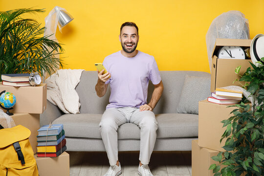 Cheerful young owner man using mobile cell phone typing sms message sit in room on sofa at home unpacking stuff indoor plant rents flat isolated on yellow wall. Relocation moving in apartment concept.