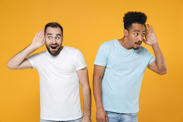 Curious young two friends european african american men 20s in white blue casual t-shirts try to hear you overhear listen intently looking aside isolated on yellow colour background studio portrait.