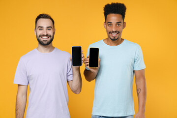 Smiling young two friends european african american men 20s in violet blue casual t-shirts using mobile cell phone with blank empty screen isolated on bright yellow colour background studio portrait.