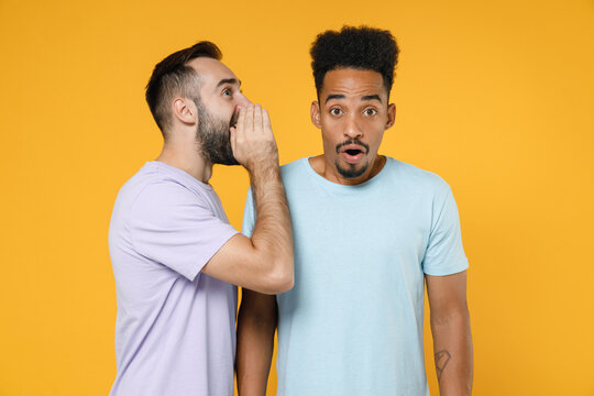 Shocked young friends european african american men 20s in casual violet blue t-shirts whispers gossip and tells secret with hand gesture isolated on bright yellow colour background studio portrait.