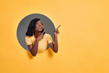 Young  woman in a round hole in the orange wall pointing and looking side. Copy space.
